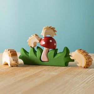 A red mushroom surrounded by green grass and three wooden hedgehog toys on a light wood surface.