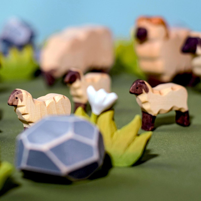 Artistic wooden toys scene with sheep in a meadow, featuring rocks, greenery, and a prominent hexagonal rock in the foreground.