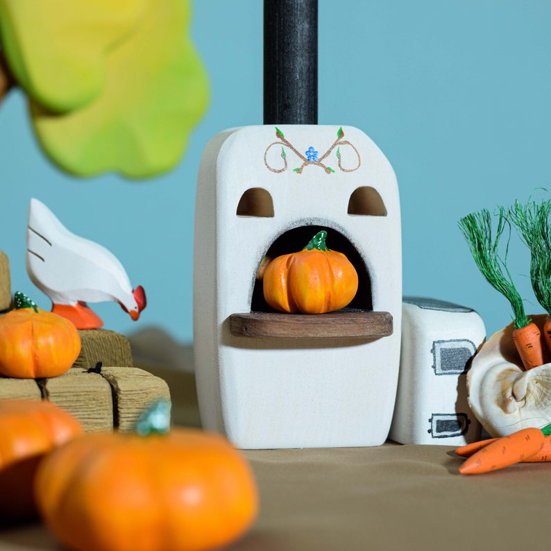 A rustic wooden stove toy with an open oven containing a pumpkin, accompanied by a hen, on a tabletop with vegetables and a wooden fence in the background.