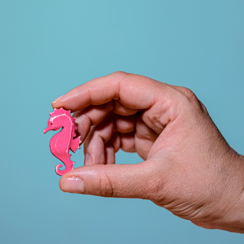 Hand holding a pink wooden seahorse figurine with a blue background, showcasing the size and craft of the piece.