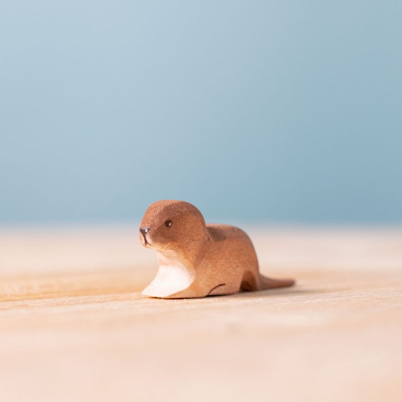 Close-up of a wooden otter figurine with a prominent, curved tail and a gentle facial expression, against a soft blue background.