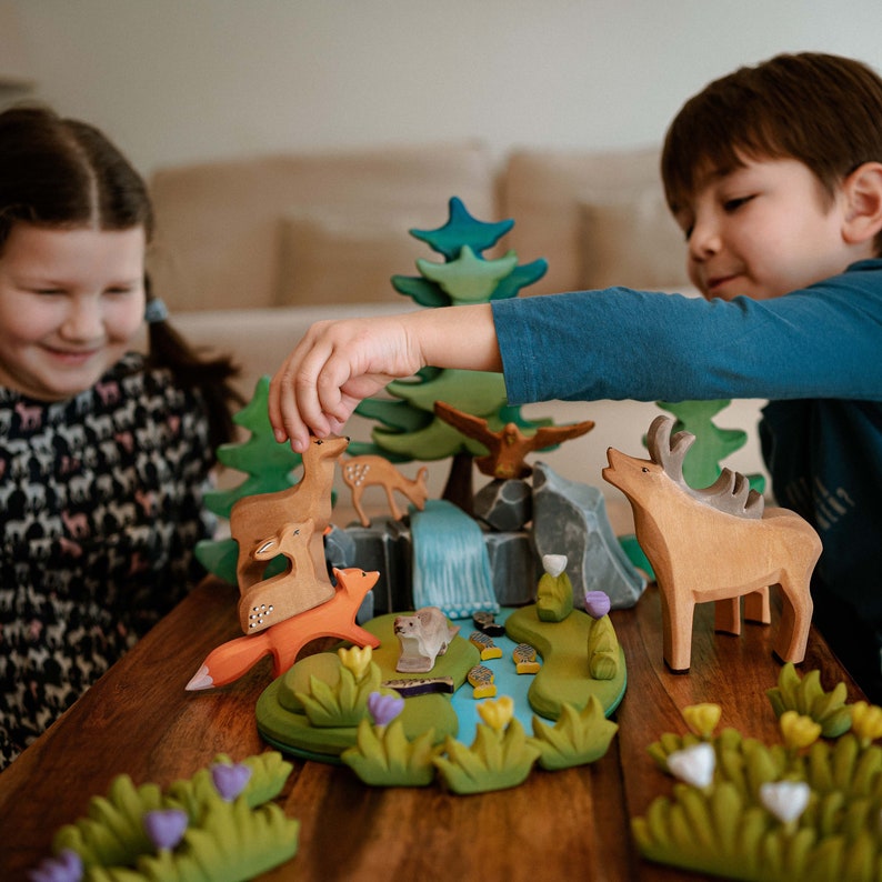 A boy and a girl smiling and playing with a variety of wooden animal figures and trees on a table, showcasing interactive playtime.