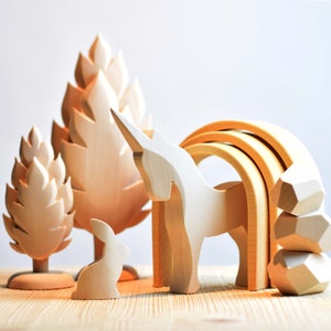 May include: Wooden toy set featuring a unicorn, a rabbit, two trees, a rainbow arch, and stacked geometric shapes. The toys are all made of natural wood and have a light brown color.