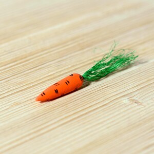 A miniature toy carrot with detailed black lines and a green top, on a natural wood surface.