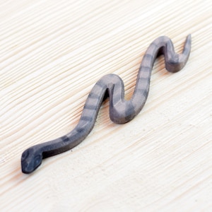 Puede incluir: Figura de serpiente de madera gris con un patrón de rayas en el cuerpo. La serpiente está tallada en forma de serpentina ondulada y descansa sobre una superficie de madera clara.