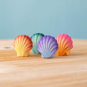 Op de afbeelding: Vier kleurrijke houten schelpenbeeldjes. De schelpen zijn beschilderd in tinten oranje, geel, groen, paars en roze. Ze zijn gerangschikt op een lichtbruin houten oppervlak met een lichtblauwe achtergrond.