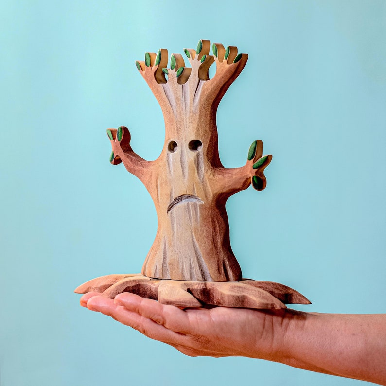 A carved wooden spooky tree supported by an open hand, showcasing the toy's size and intricate details, against a calm blue background.