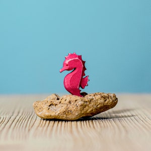 A vibrant pink wooden seahorse figurine perched on a rocky base against a serene blue background, evoking a whimsical oceanic theme.