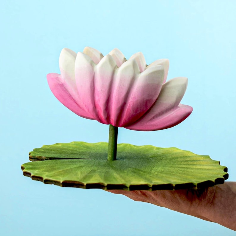 A hand holds a wooden lotus flower with soft pink petals and a bright green lily pad base against a gentle blue background.