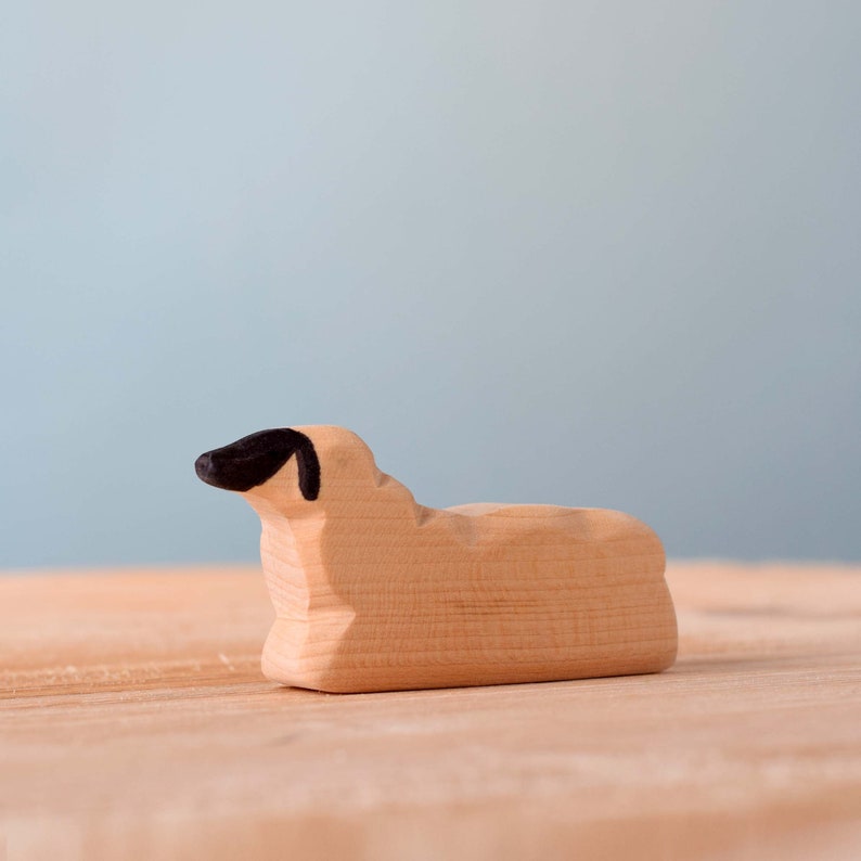 A wooden sheep toy lying down on a wooden surface, with detailed painting on its face to represent its eyes and nose.