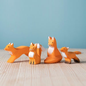 Family of four wooden fox toys with orange and white detailing, standing on a wooden surface against a light blue background.