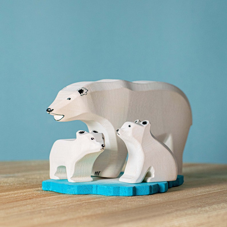 A family of wooden polar bear figurines consisting of a large bear and two cubs on a blue painted wooden base, against a pale blue backdrop.