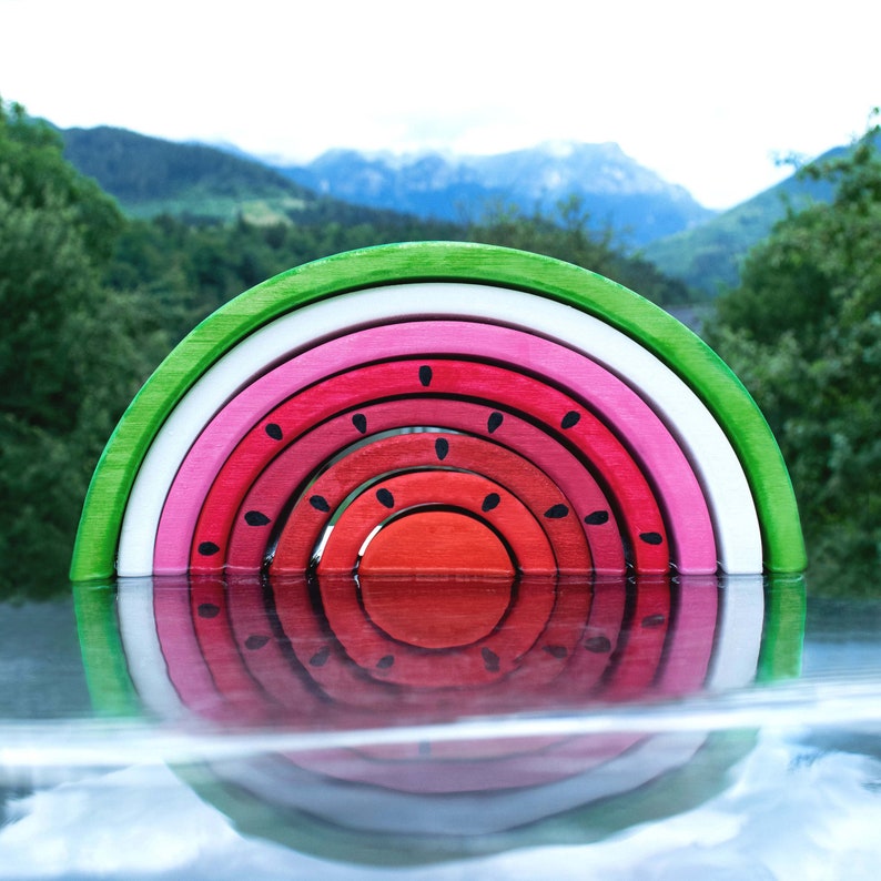 Wooden watermelon stacker toy against a mountainous backdrop, reflecting in water, nature-inspired Montessori toy.