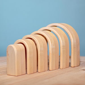 May include: A set of seven natural wooden rainbow arches, arranged in size order from smallest to largest, on a light brown wooden surface.