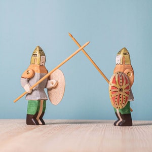 Two wooden knight figurines with lances, shields, and a medieval theme, facing each other as if in a friendly duel, on a wooden surface with a pale blue background.