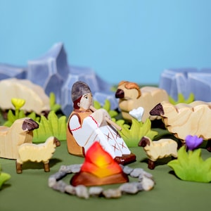 A wooden figurine of a person in traditional dress seated by a fire, surrounded by sheep and lush greenery on a crafted landscape.