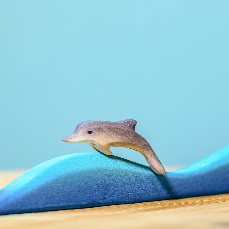 A lone wooden dolphin, part of BumbuToys' marine collection, is poised mid-jump above a soft blue wave, evoking the playful essence of ocean life against a pale blue setting.