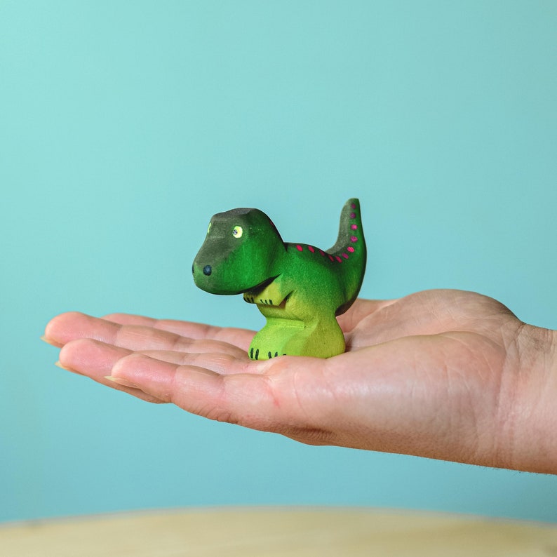 Human hand holding a small wooden T-Rex toy, showcasing the toy's size, with green body and magenta back spots.