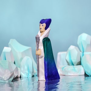 A wooden figure with a purple head, white robe, and green cloak holding a silver staff among icy blue wooden formations.