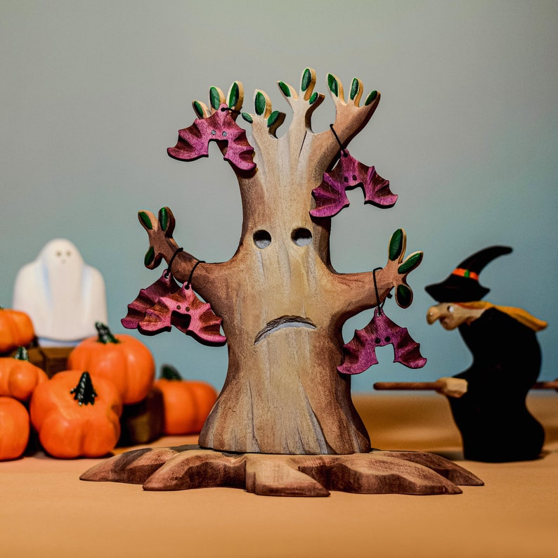 Artisanal wooden tree toy with multiple hanging bat figurines, accompanied by a wooden ghost and pumpkins against a soft backdrop.