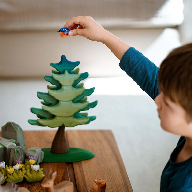 A child reaching to place a wooden kingfisher toy atop a green wooden tree, embodying BumbuToys’ philosophy of hands-on learning and development of fine motor skills through play.