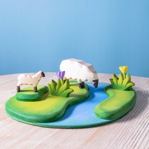 Handcrafted wooden sheep on a painted play mat with green grass and blue water details, with bushes and flowers.