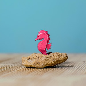 The pink wooden seahorse figurine from a different angle, emphasizing the playful contrast between the figure and the soft blue background.