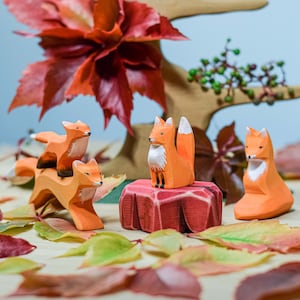 Group of wooden fox toys surrounded by autumn leaves, with a red tree backdrop, creating a seasonal forest scene.