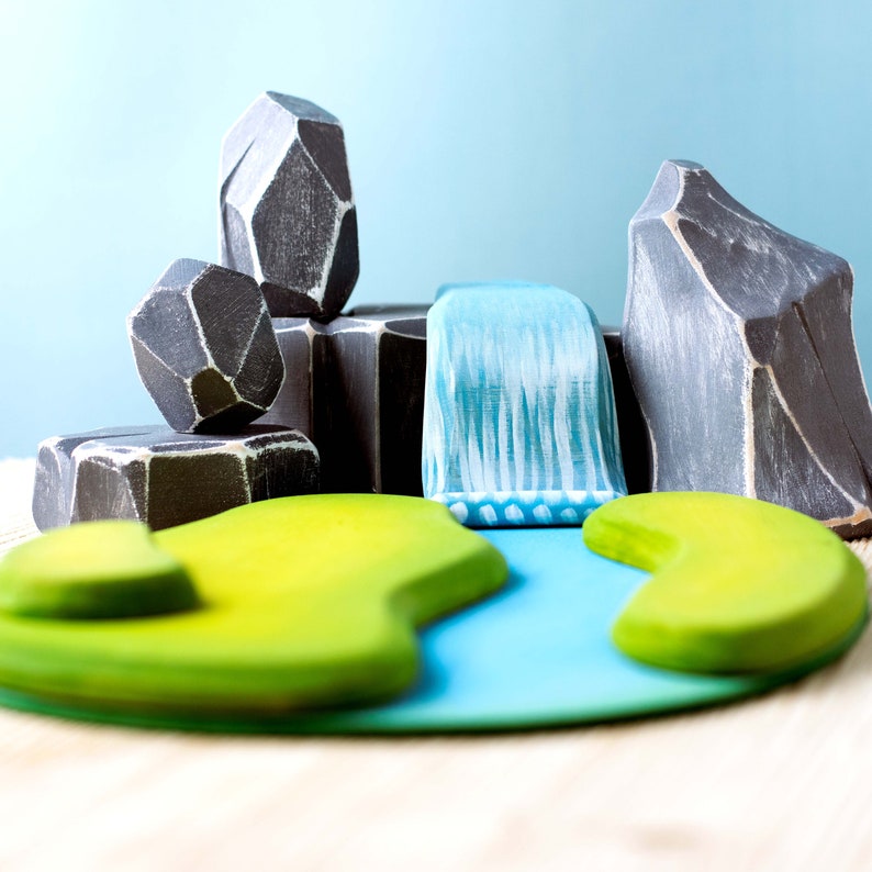 Colorful wooden playset featuring grey mountains, a blue waterfall, and green island pieces on a wooden surface against a light blue background.