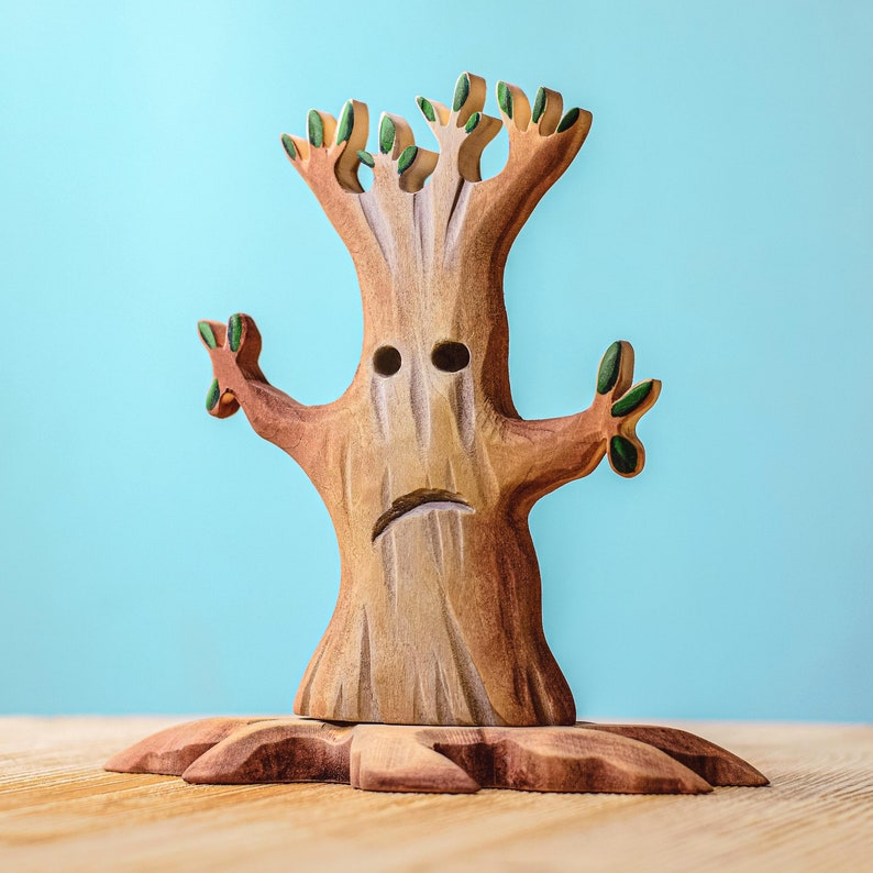 Front-facing image of a wooden spooky tree with a forlorn expression and lush green leaves, standing tall on a wooden base against a serene blue backdrop.