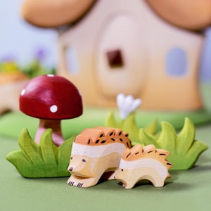 Two wooden hedgehog toys near a red mushroom in a whimsical setting with a wooden house backdrop on a green base.