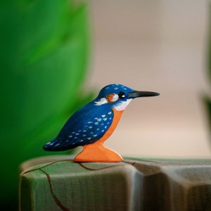 Artisanal wooden kingfisher figurine painted in vivid blues and oranges, perched gracefully on a wooden block against a leafy green background, from BumbuToys’ handcrafted Birds Series.