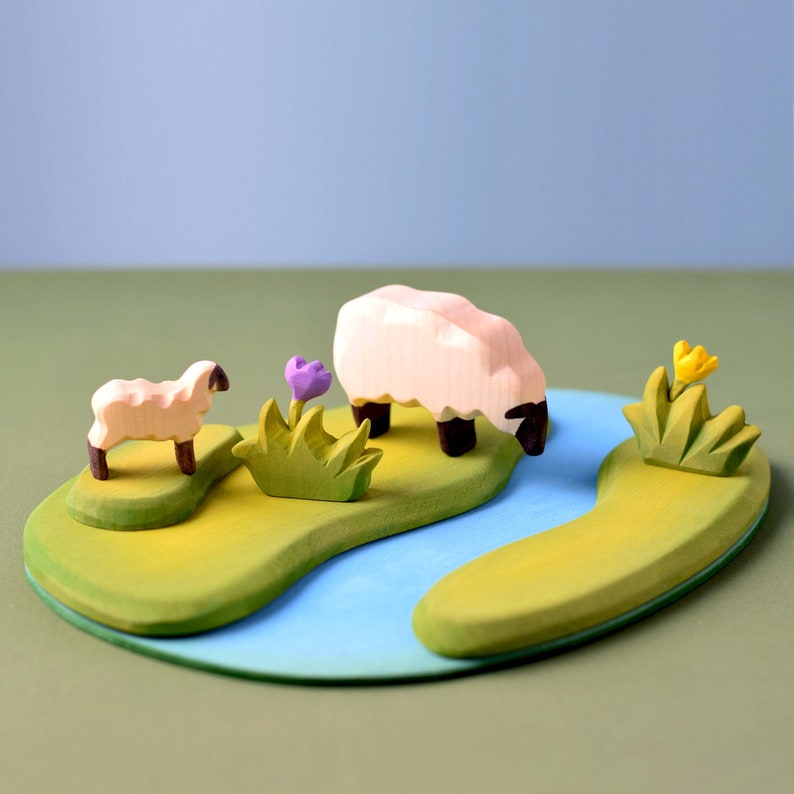 Wooden sheep toys on a painted landscape with blue rivers, green fields, and purple flowers against a pale blue background.