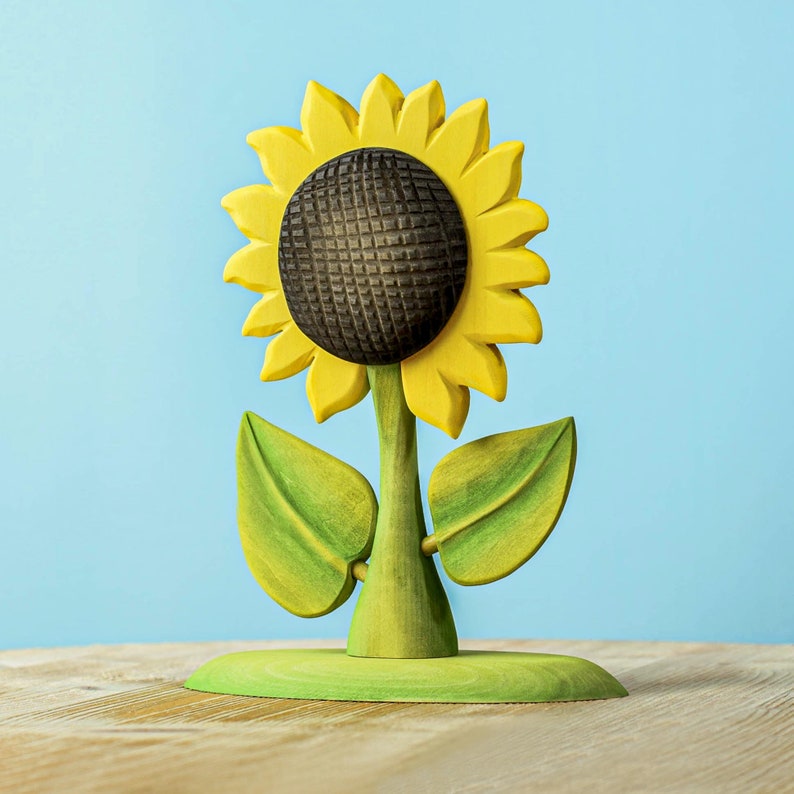 Wooden play sunflower standing with bright yellow petals and green leaves against a pale blue wall