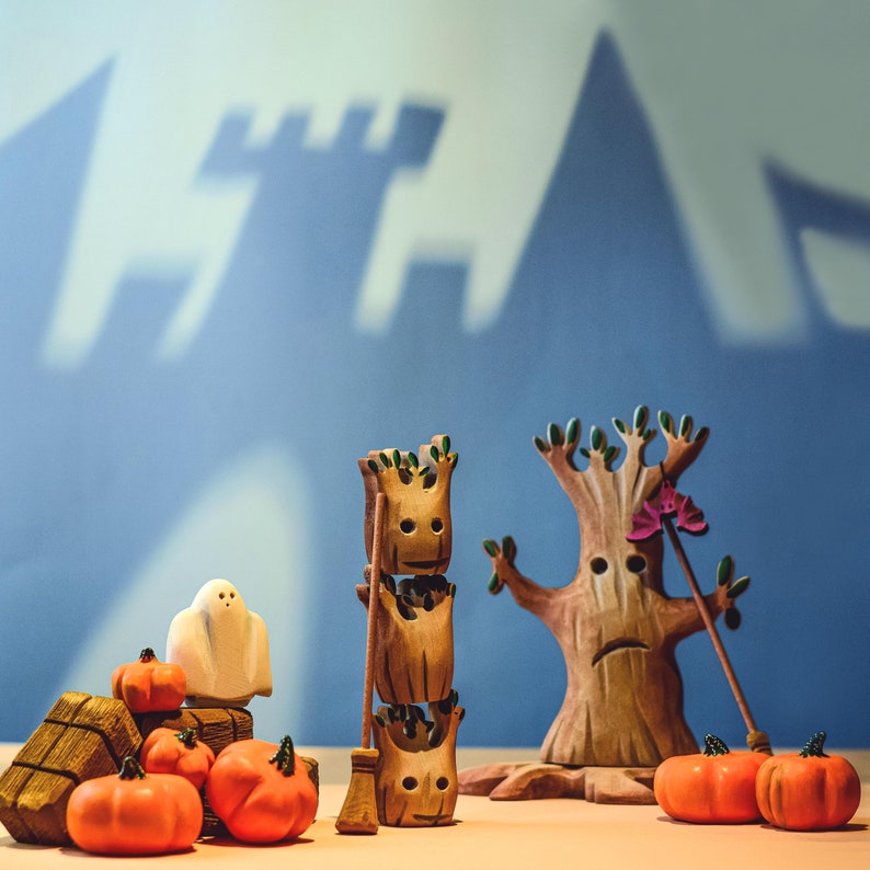 A whimsical wooden tree toy with bat figurines, flanked by a ghost and pumpkins, casting a silhouette on a blue backdrop with castle outlines.