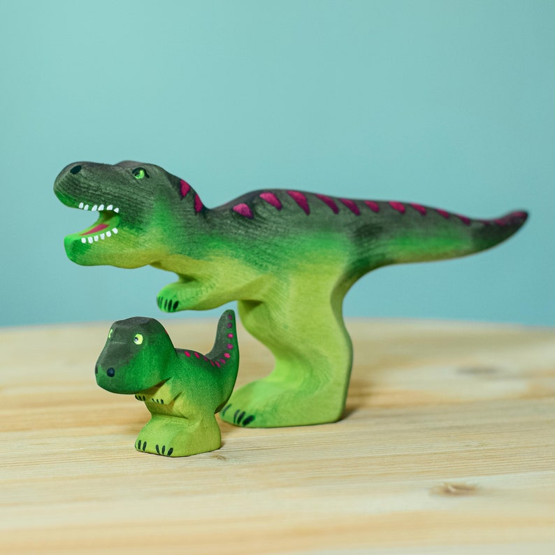 Handcrafted wooden T-Rex toy in mid-roar with vibrant green body and magenta details, next to its smaller counterpart on a wooden surface.