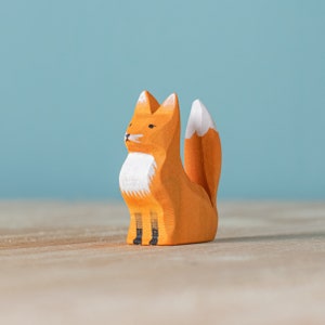 Wooden fox toy seated upright with white detailing on the face and tail, on a wooden surface against a pale blue background.