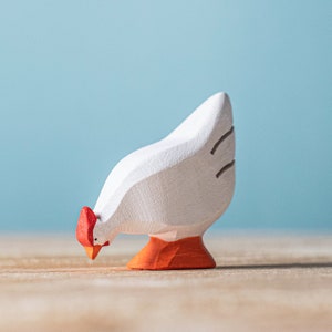 A hand-painted wooden hen toy with a white body, red comb, and orange feet, pecking at the ground on a wooden surface with a light blue backdrop.