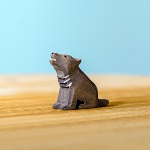 Hand-carved wooden wolf pup sitting and howling, showcasing detailed craftsmanship and natural wood patterns, on a wooden tabletop with a pale blue background.