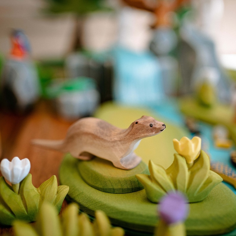 Focused view of a wooden otter figurine on a green, layered play mat with floral decorations, highlighting the toy's potential for imaginative play.