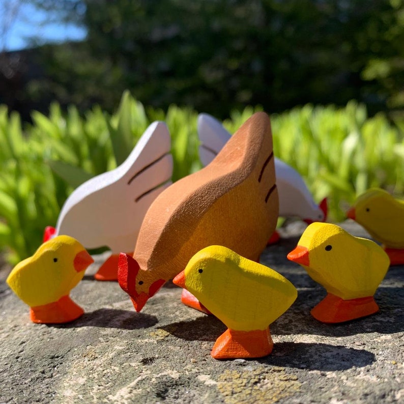 A wooden rooster toy painted in brown and white with three yellow chicks following, set on a rock outdoors with greenery in the backdrop.