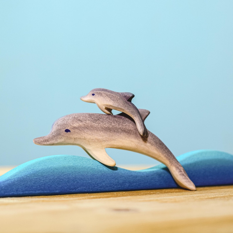 A wooden dolphin duo, elegantly painted, demonstrates the beauty of marine life through sustainable play with BumbuToys’ handcrafted aquatic series, set against a gentle blue background.