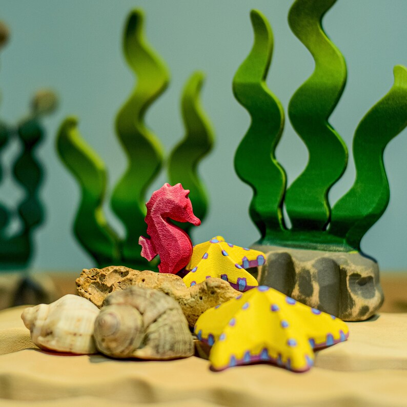 A closer look at the pink wooden seahorse figurine among textured sea plants and colorful starfish.