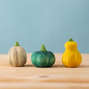 May include: Three wooden pumpkins in a row, each with a different shape and color. The first is a light tan color, the second is green, and the third is yellow. Each pumpkin has a green stem with white dots.
