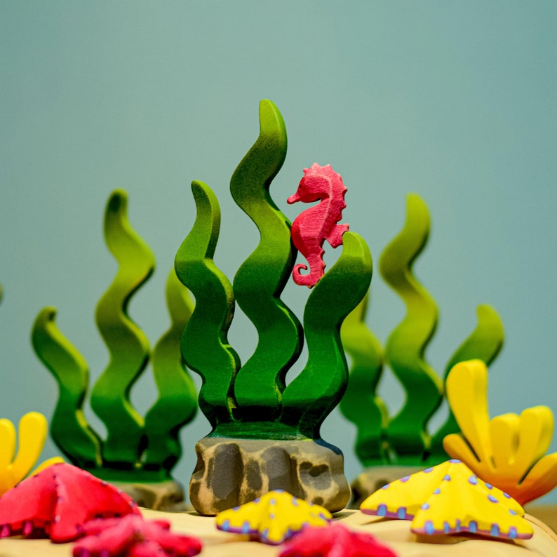 A closer look at the pink wooden seahorse figurine among textured sea plants and colorful starfish, highlighting the details of the wooden craftsmanship.