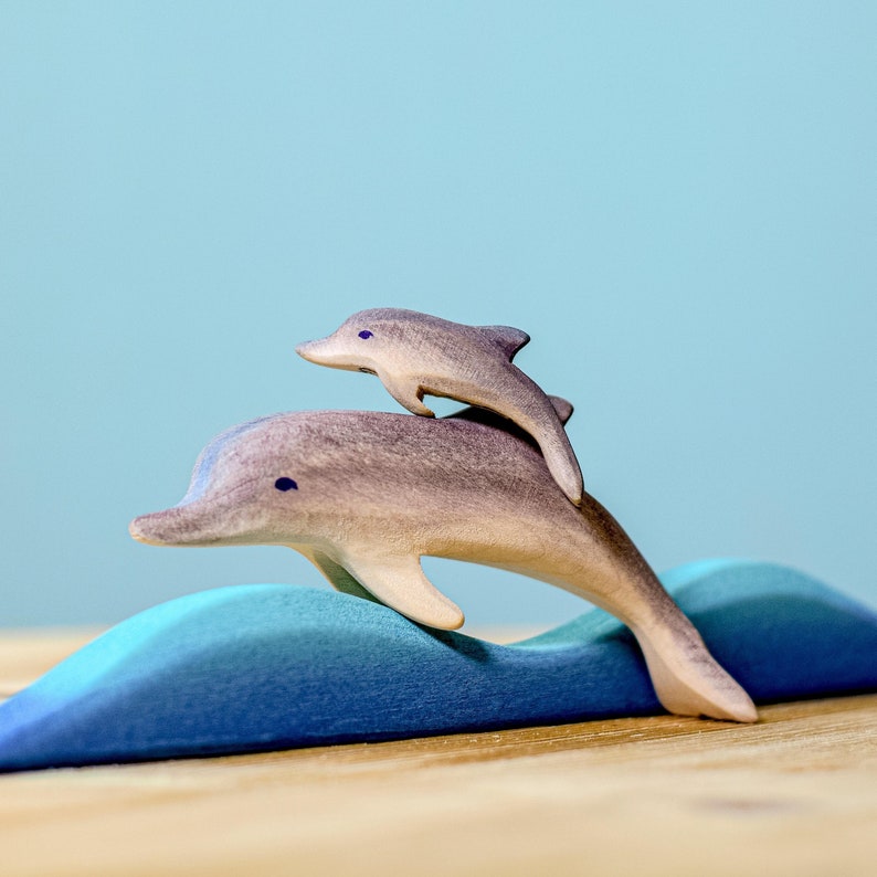 Two wooden dolphins, masterfully crafted, leap together above stylized blue waves, captured against a serene blue backdrop, highlighting the playful spirit and intricate artisanship synonymous with BumbuToys.