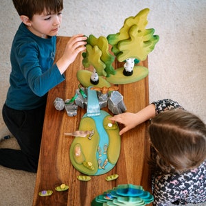 Two children engaged in creative play with a wooden river and landscape set, including trees and animals, on a brown table.