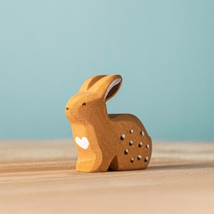 A small, handmade wooden bunny figurine with decorative details stands against a blue background, evoking a serene and simplistic charm.
