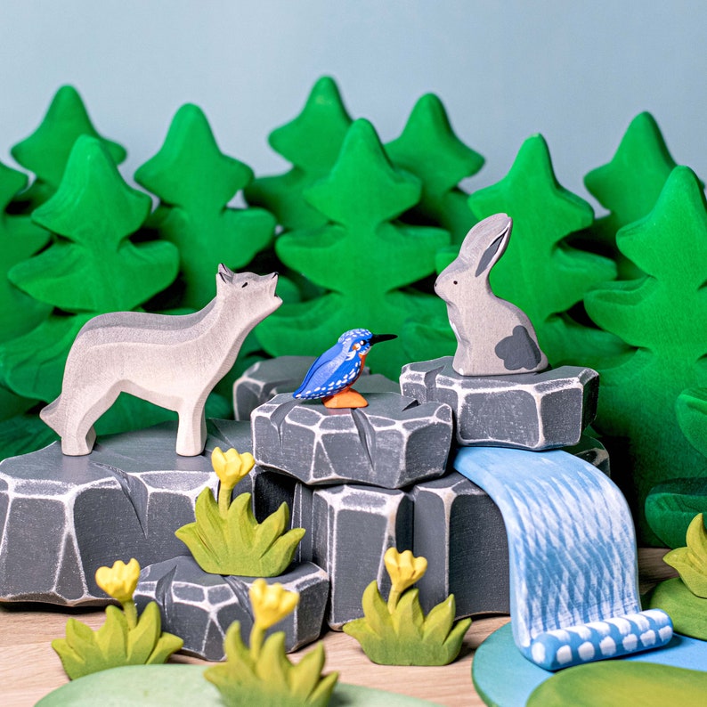 Scene with wooden wolf, rabbit, and bird figurines among green trees and grey rocks with a blue river.