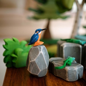 Brightly colored wooden kingfisher toy on a stone, surrounded by wooden greenery, showcasing BumbuToys’ dedication to crafting natural and imaginative play settings.